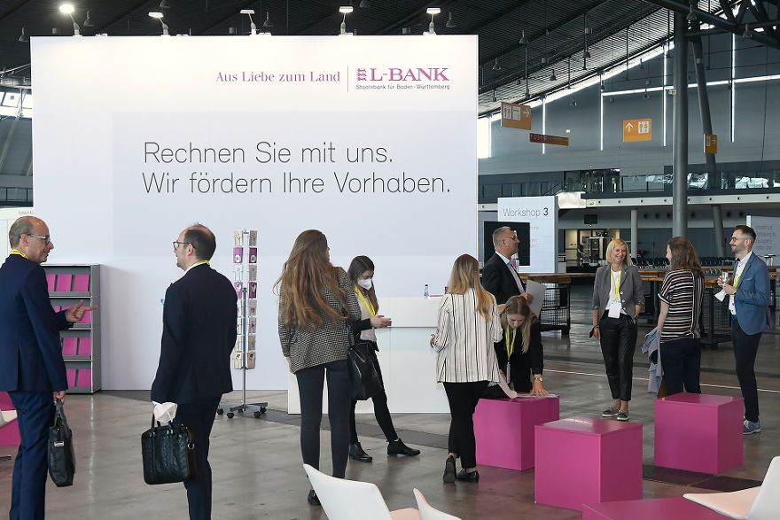 Mehrere Menschen stehen in Gruppen zusammen und unterhalten sich, im Hintergrund steht ein Banner mit der Aufschrift Rechnen Sie mit uns. Wir fördern Ihre Vorhaben. und dem Logo der L-Bank.