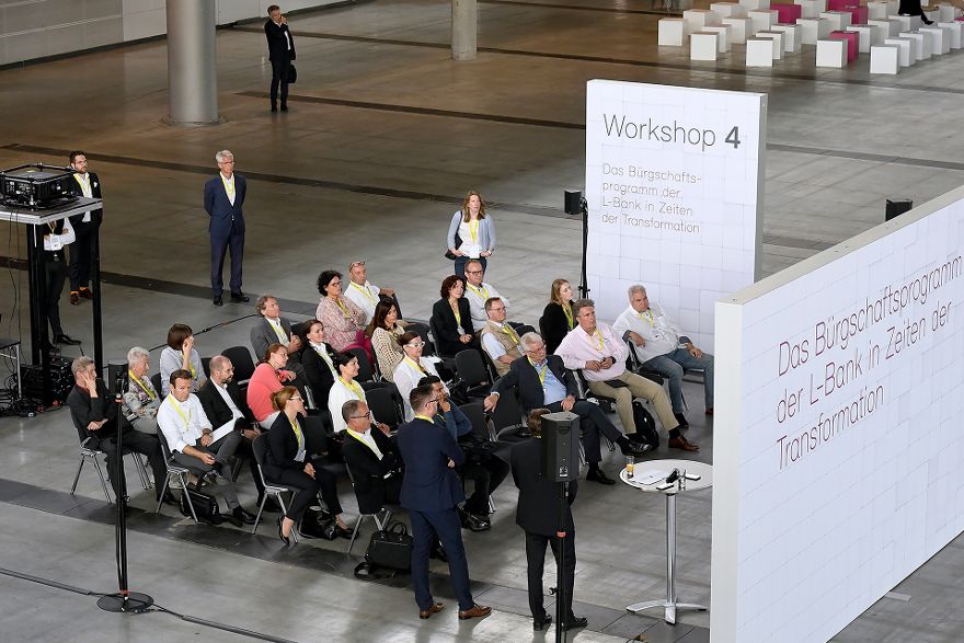 Eine Gruppe an Menschen sitzt in vier Stuhlreihen zusammen und schaut nach vorne, im Hintergrund steht ein Banner mit der Aufschrift Workshop 4/Das Bürgschaftsprogramm der L-Bank in Zeiten der Transformation