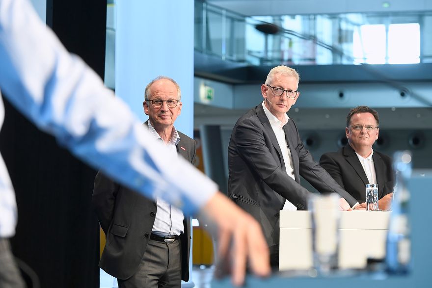 Rainer Reichhold, Moderator Joachim Dorfs und Claus Paal stehen auf einer Bühne, im Vordergrund ist unscharf der Arm einer weiteren Person zu sehen.