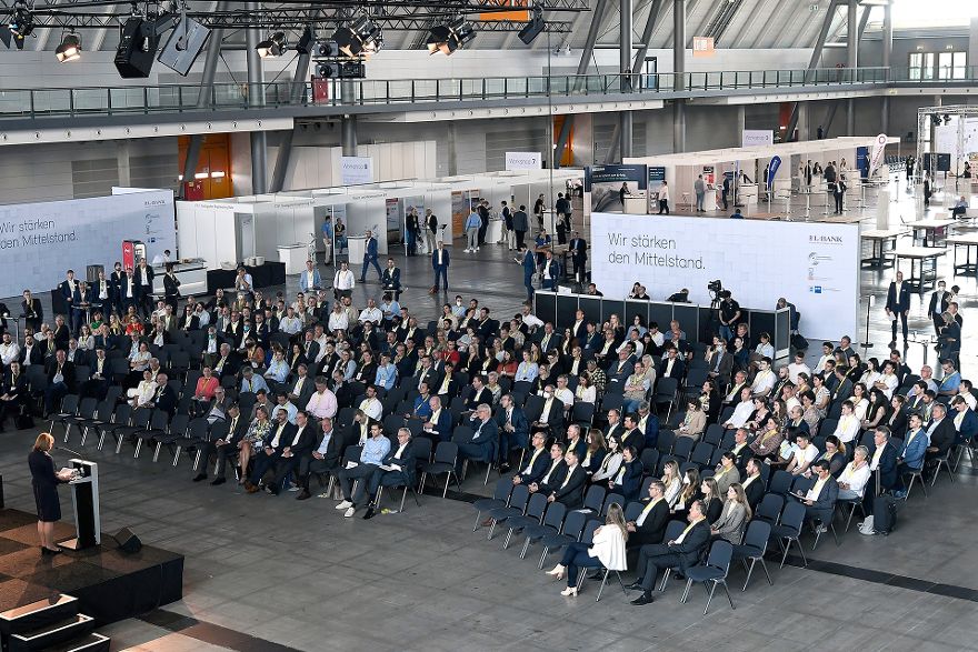 Blick von schräg oben auf ein Publikum in mehreren Sitzreihen, das auf eine Bühne unten links schaut, auf der Edith Weymayr am Rednerpult steht.