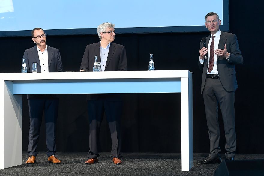 Guy Selbherr, Moderator Klaus Köster und Johannes Heinloth stehen hinter einen Tisch auf der Bühne.