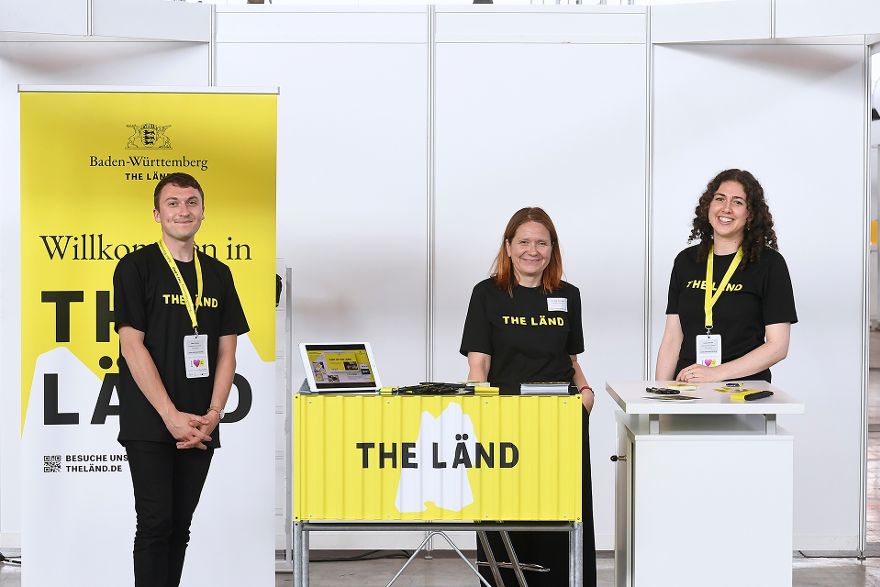 Ein Mann und zwei Frauen stehen an einem Stand und schauen in die Kamera, der Schriftzug The Länd ist zu sehen.