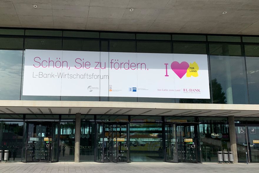 Eingangsbereich einer Halle, über den Drehtüren hängt ein Banner mit der Aufschrift Schön Sie zu fördern. L-Bank-Wirtschaftsforum.
