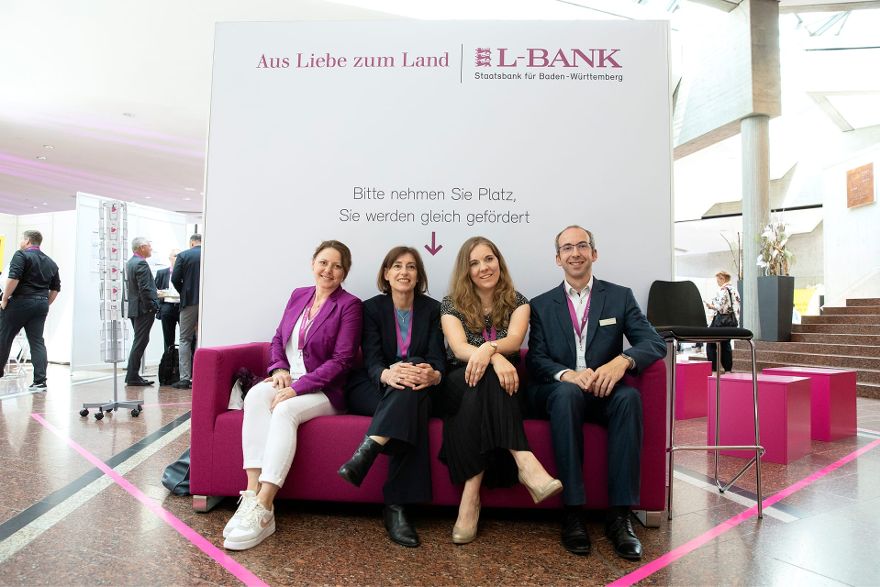 Vier Personen sitzen auf einer magentafarbenen Couch, dahinter steht ein Banner mit der Aufschrift Bitte nehmen Sie Platz, Sie werden gleich gefördert. Darüber ist das Logo der L-Bank zu sehen.
