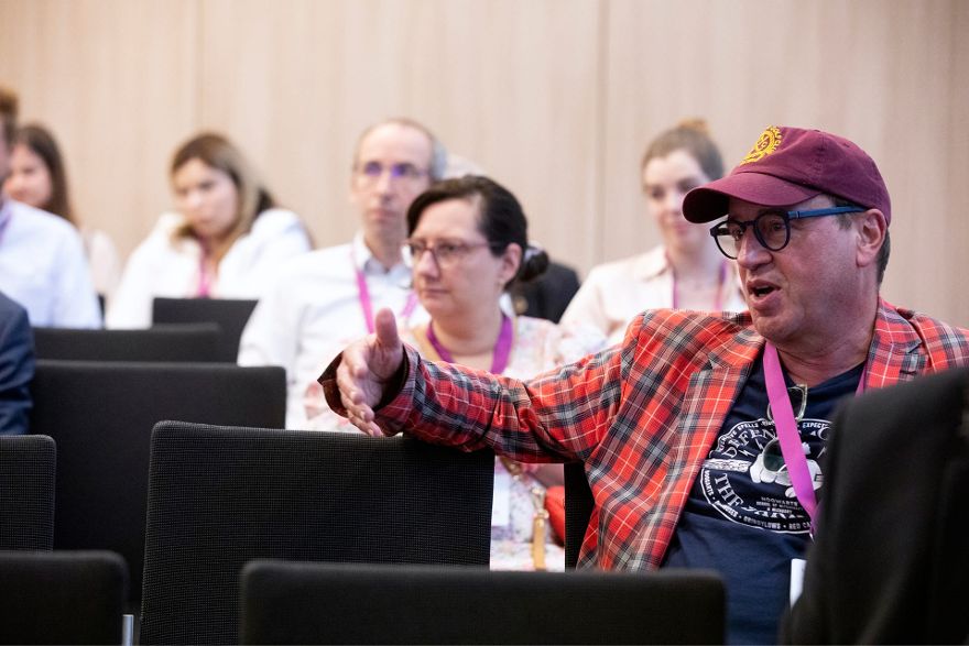 Ein sitzender Mann mittleren Alters mit Hemd, Brille und Baseball-Mütze redet, hinter ihm sitzen weitere Personen.