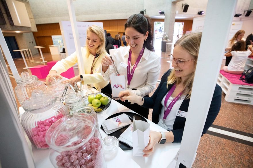 Drei Frauen stehen lächelnd hinter einem Tisch, auf dem Gläser mit Bonbons und eine Schale Obst stehen. Mithilfe von Greifzangen füllen sie Tütchen mit den Bonbons.